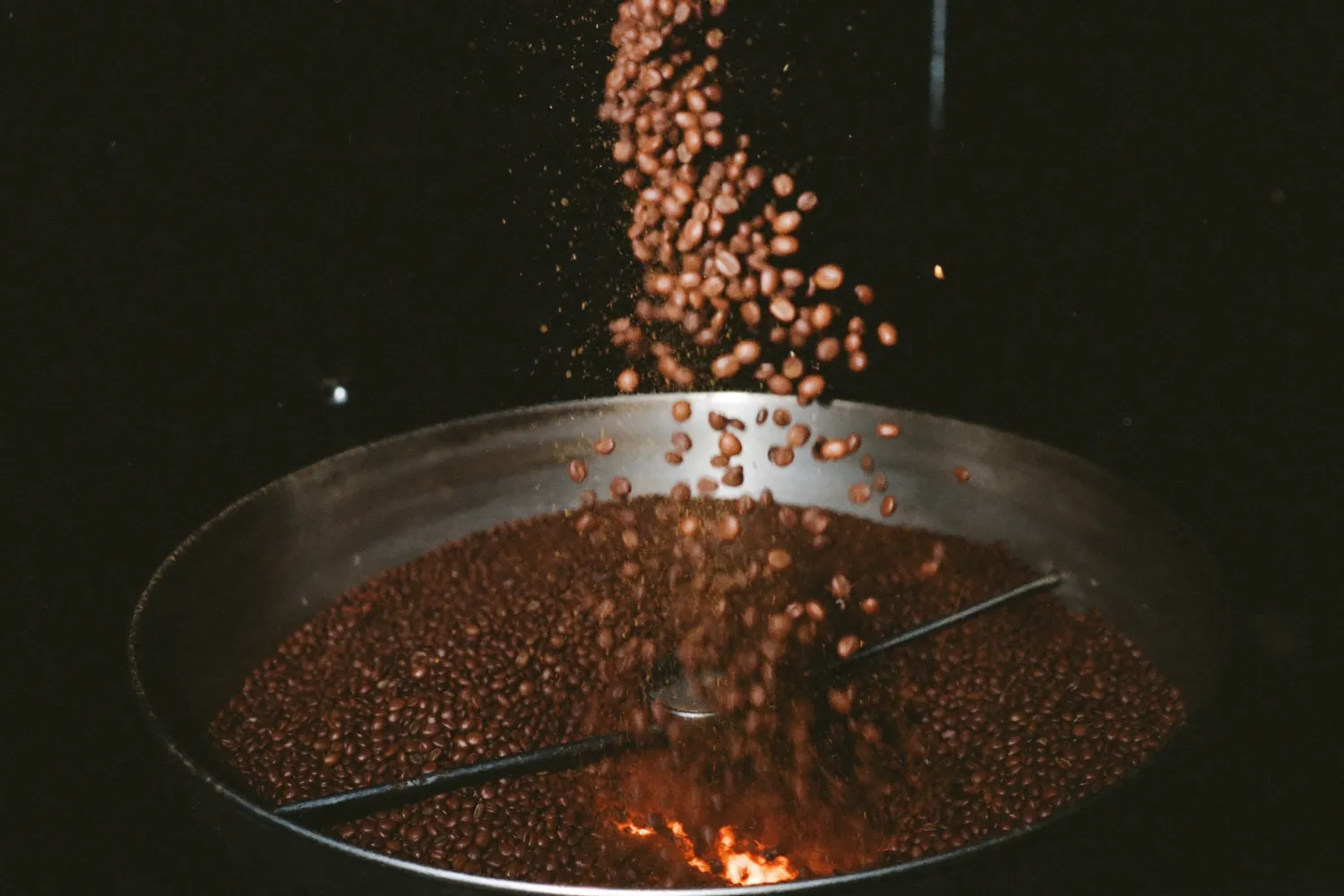 Grãos de café em cascata dentro do tambor quente, iluminados por brasa.