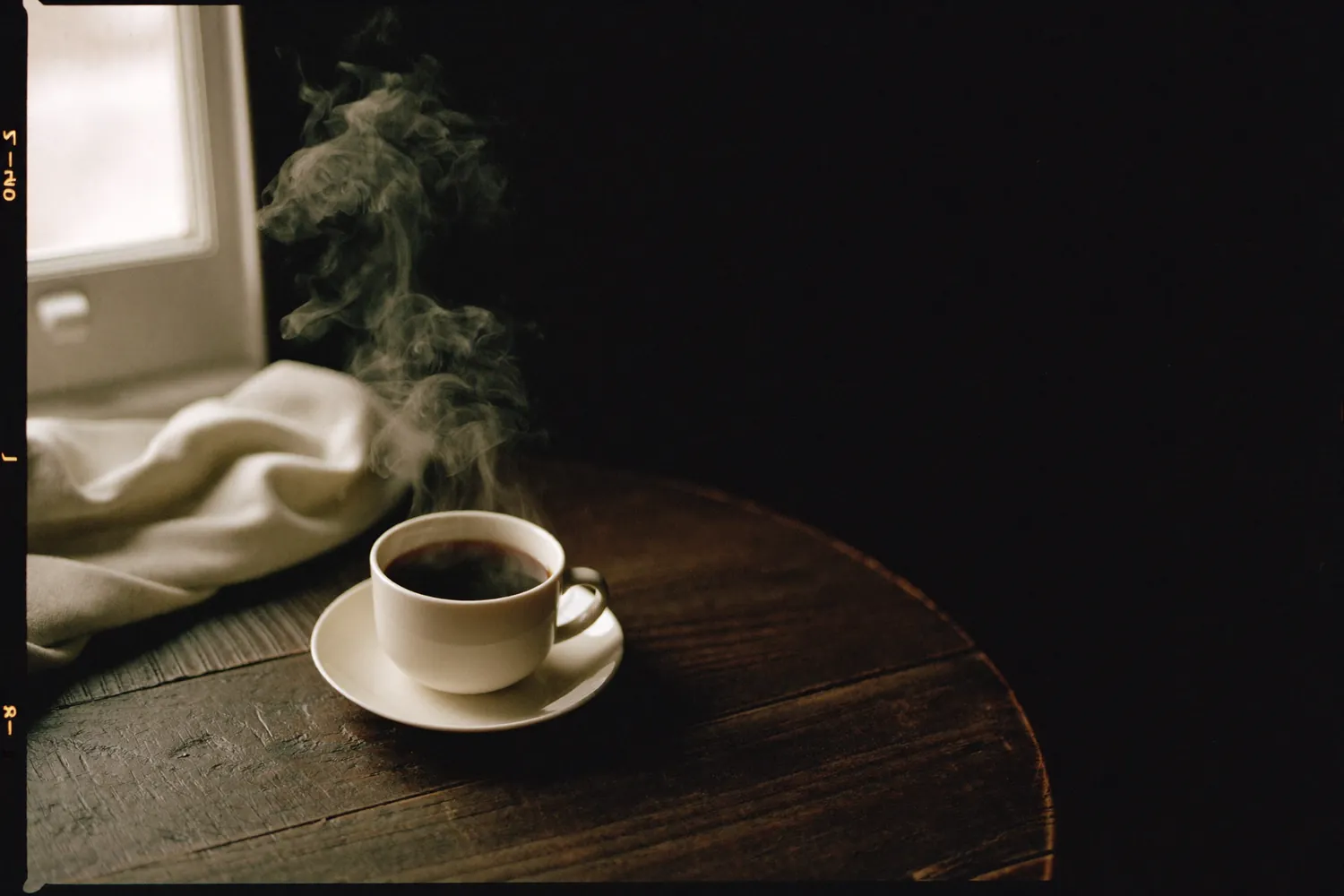 Xícara branca de café preto sobre mesa de madeira escura, com vapor.